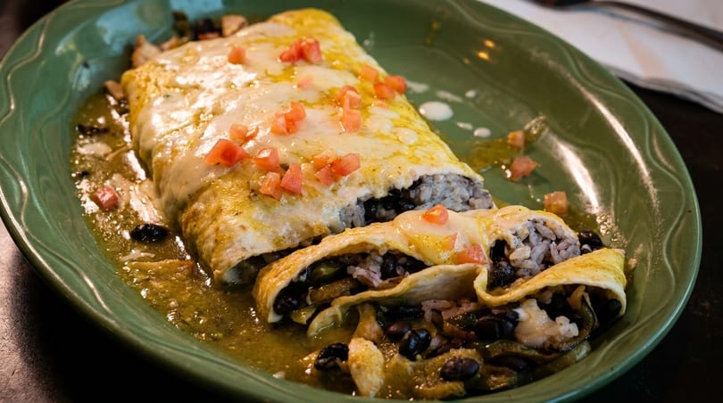 Two cheesy enchiladas with rice, black beans, and green sauce, topped with diced tomatoes.
