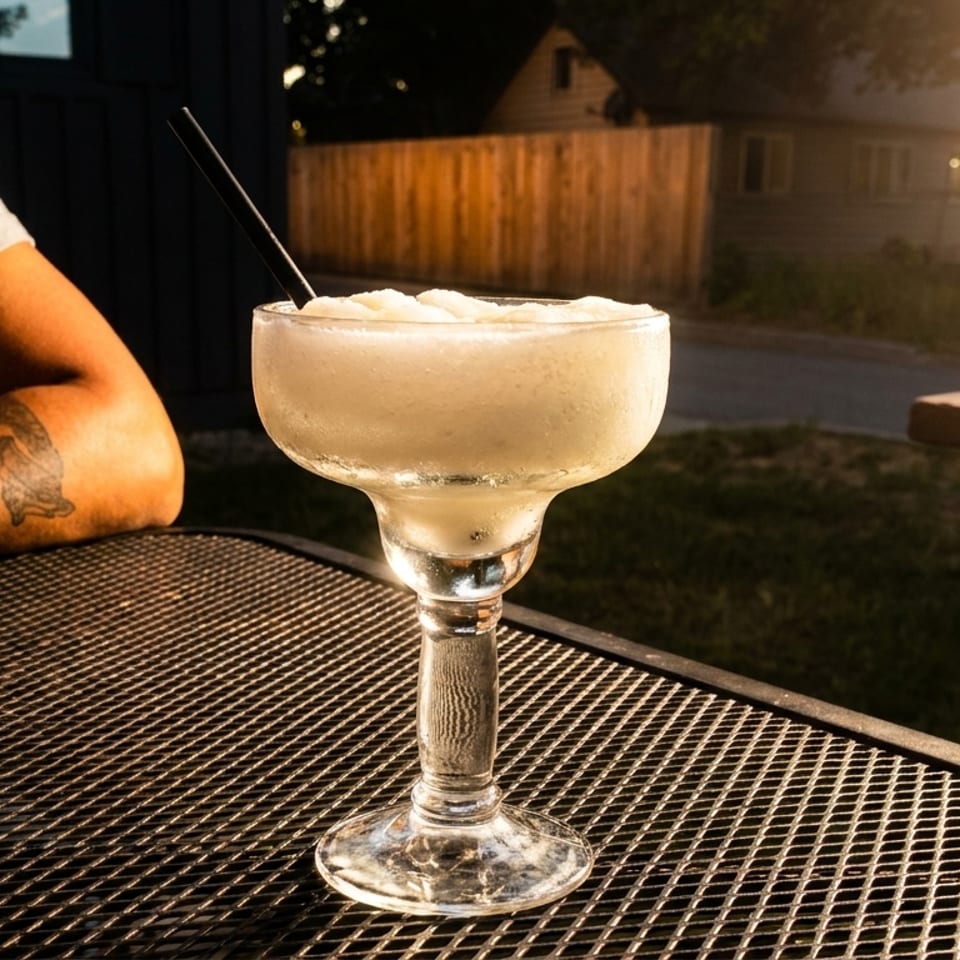 Frozen margarita on a patio table with a person's arm and a sunny background.