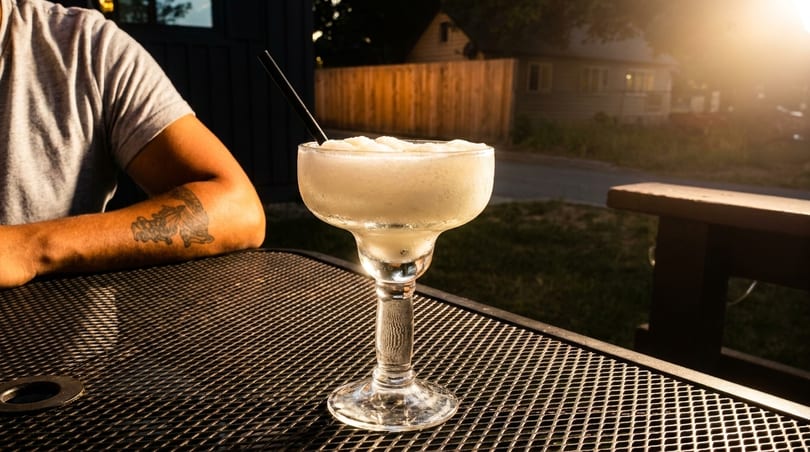 Frozen margarita on a patio table with a person's arm and a sunny background.