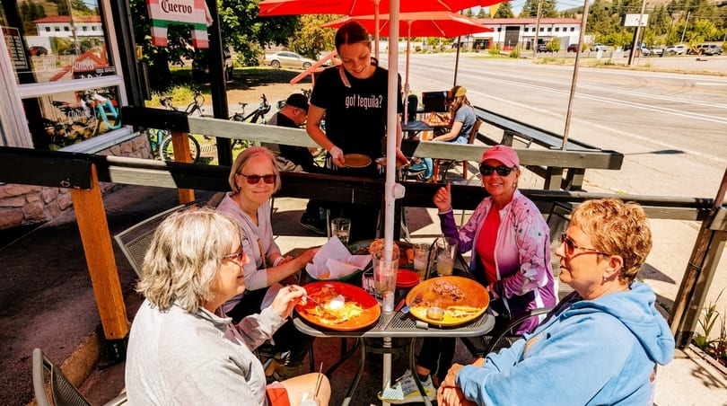 Four women dining outdoors at a restaurant, with a server delivering food to their table.